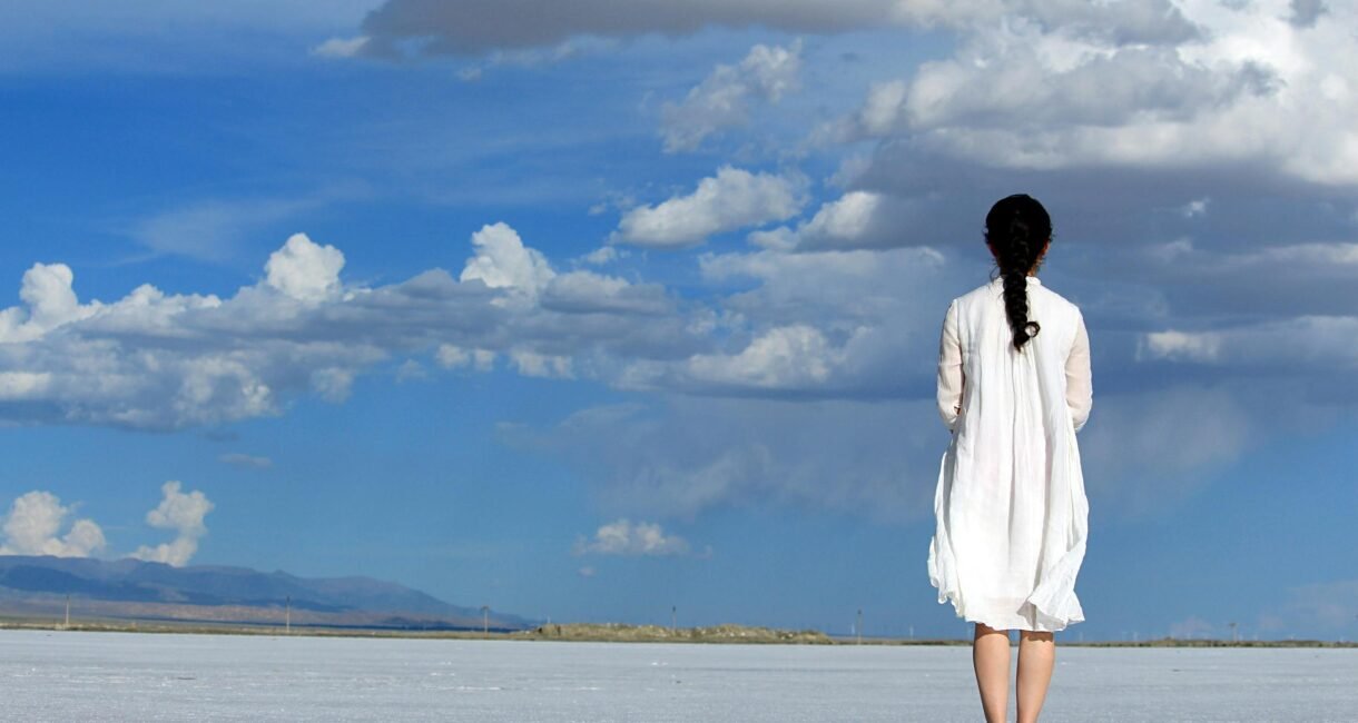Woman stands alone on a vast salt flat under a bright blue sky with clouds.