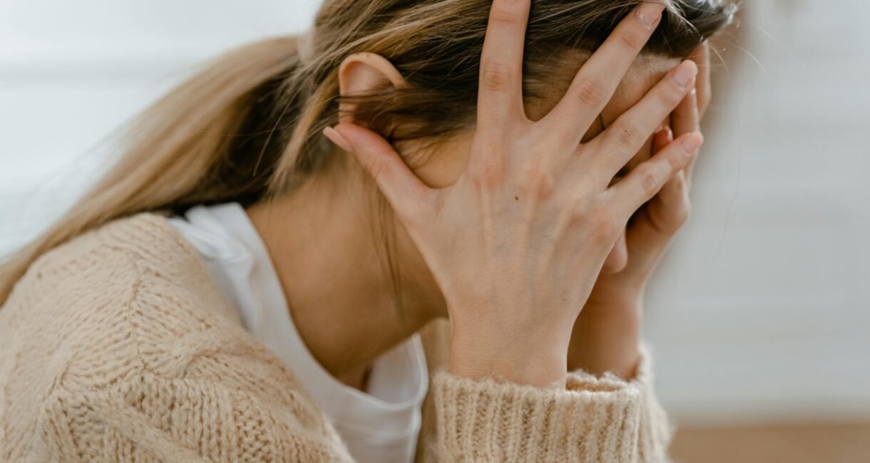 Woman sitting indoors with face covered by hands, expressing stress and frustration.