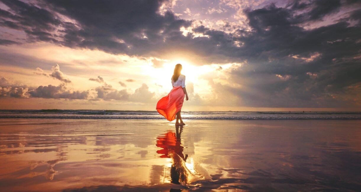 A woman stands on a reflective beach at sunset, wearing a flowing skirt, under a dramatic sky.