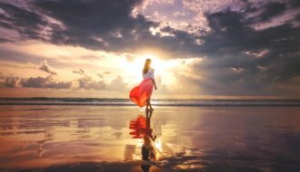 A woman stands on a reflective beach at sunset, wearing a flowing skirt, under a dramatic sky.