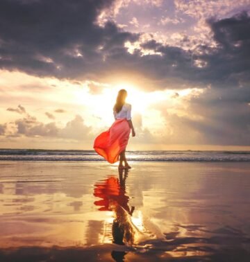 A woman stands on a reflective beach at sunset, wearing a flowing skirt, under a dramatic sky.