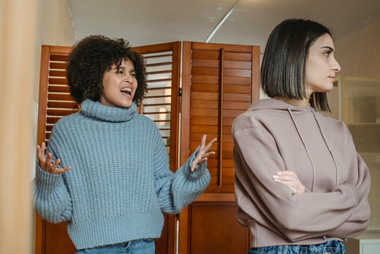 Furious African American female with black hair screaming while fighting with displeased female in light room with wooden folding screen