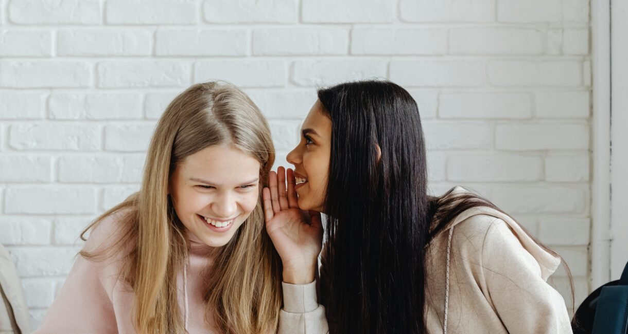 Two young women sharing a secret and laughing, dressed casually, indoors.