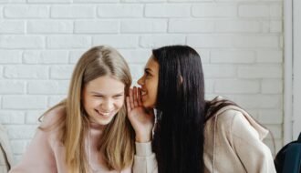 Two young women sharing a secret and laughing, dressed casually, indoors.