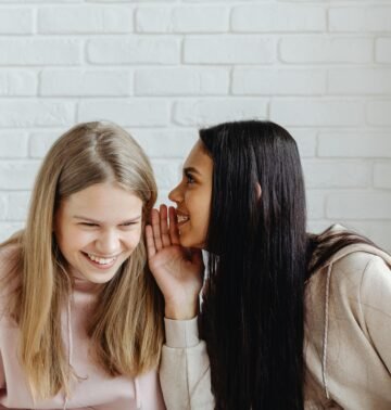Two young women sharing a secret and laughing, dressed casually, indoors.