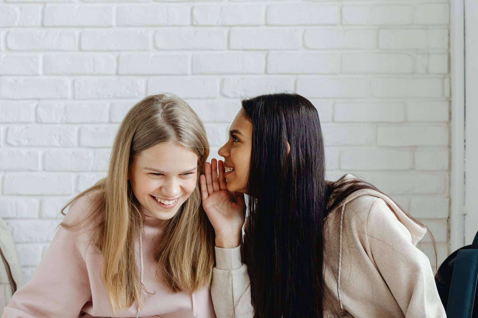 Two young women sharing a secret and laughing, dressed casually, indoors.