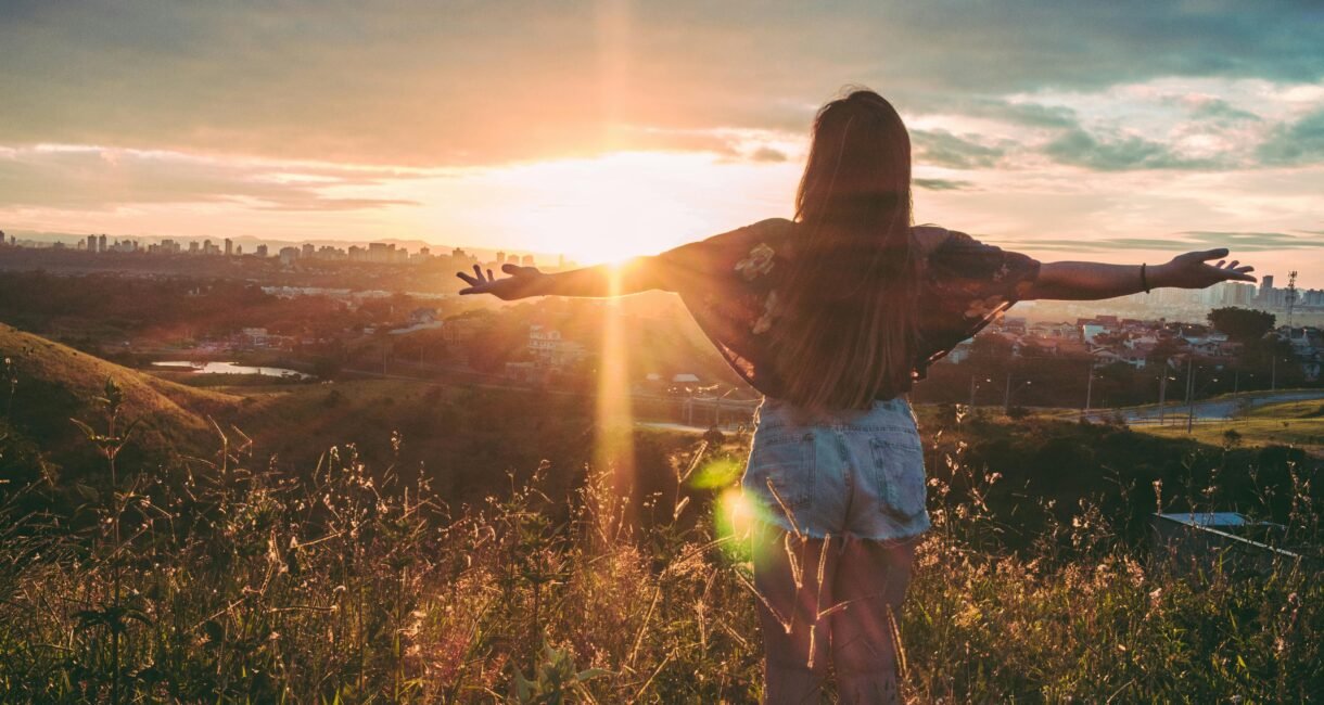 Woman with open arms enjoying sunset view over cityscape from a grassy hilltop.