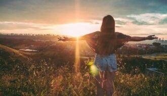 Woman with open arms enjoying sunset view over cityscape from a grassy hilltop.