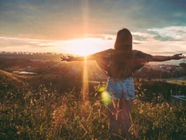 Woman with open arms enjoying sunset view over cityscape from a grassy hilltop.