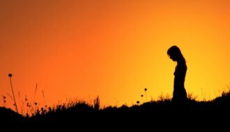 Silhouette of a woman standing in a field during a vibrant orange sunset.