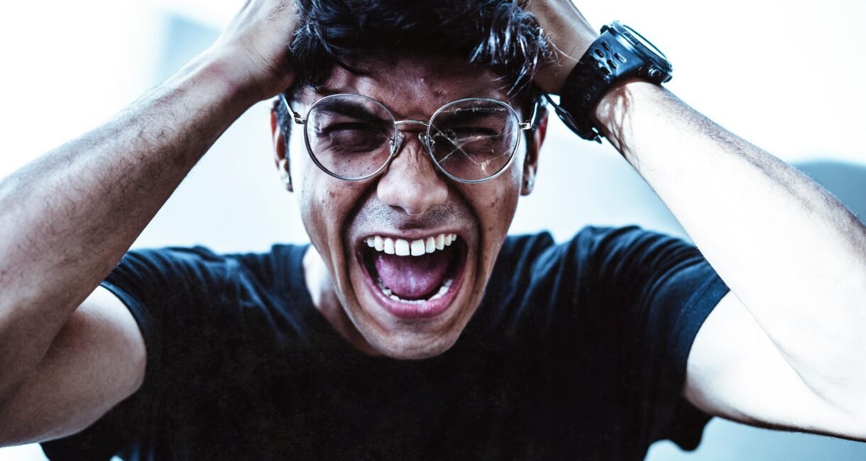 A close-up of a man shouting in frustration, clutching his head with broken eyeglasses on.