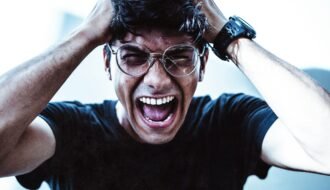 A close-up of a man shouting in frustration, clutching his head with broken eyeglasses on.