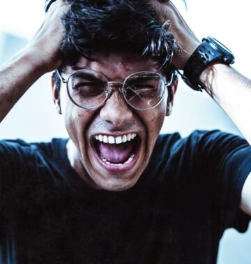 A close-up of a man shouting in frustration, clutching his head with broken eyeglasses on.