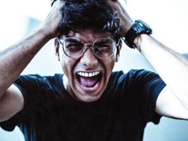 A close-up of a man shouting in frustration, clutching his head with broken eyeglasses on.