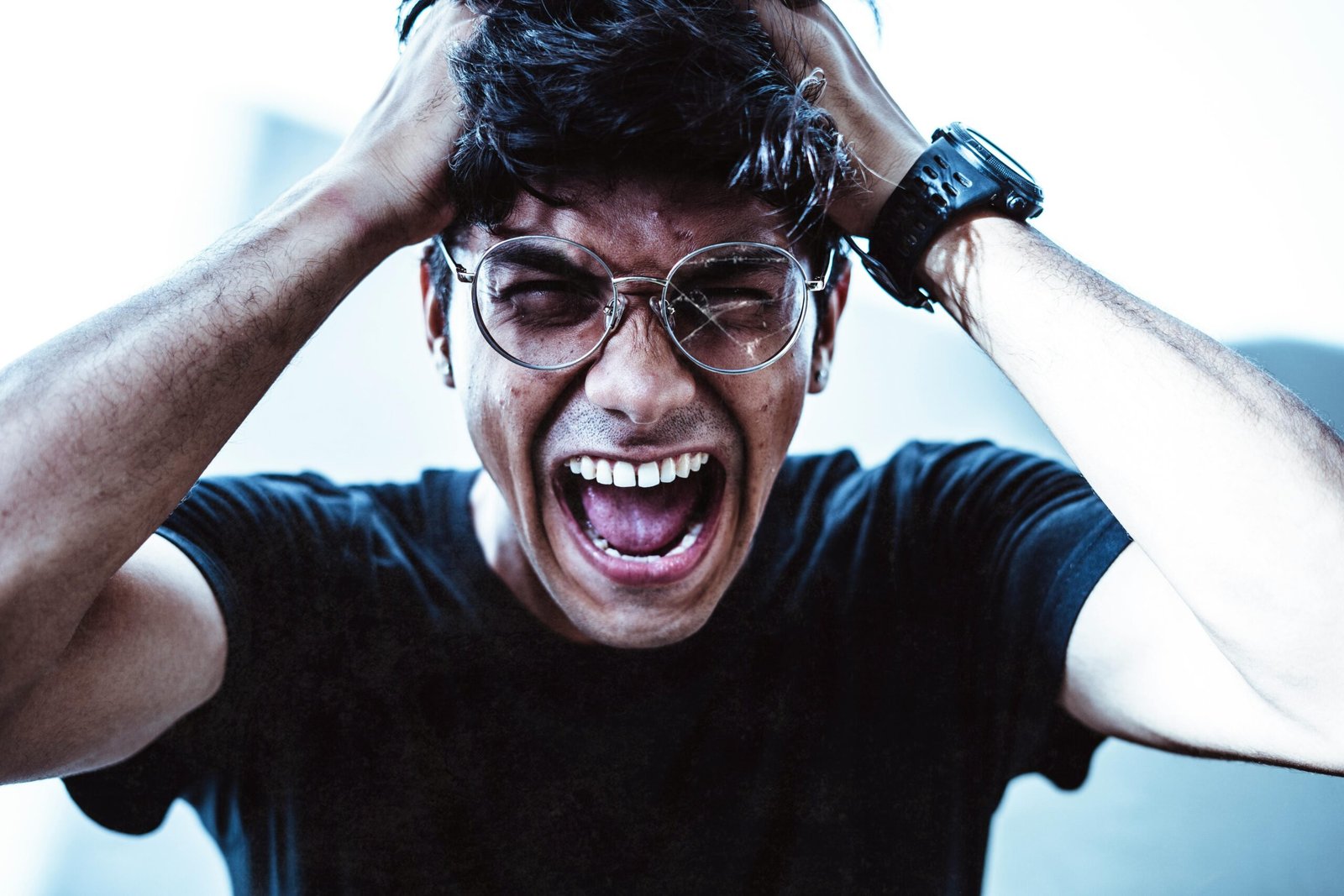 A close-up of a man shouting in frustration, clutching his head with broken eyeglasses on.