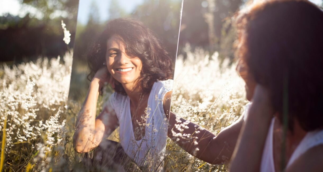 A woman smiles at her reflection in a mirror placed in a sunny field.