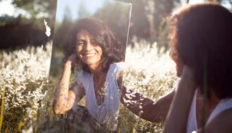 A woman smiles at her reflection in a mirror placed in a sunny field.