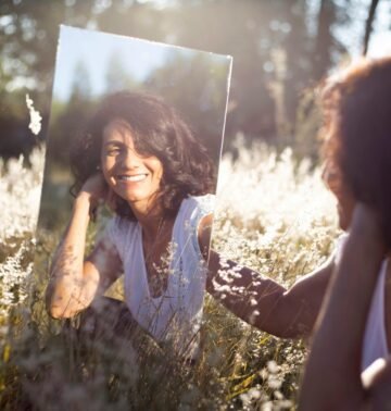 A woman smiles at her reflection in a mirror placed in a sunny field.