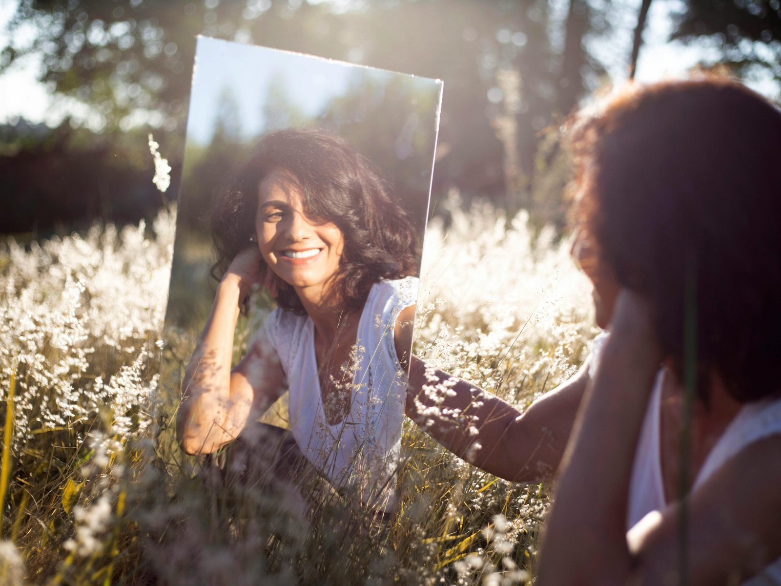 A woman smiles at her reflection in a mirror placed in a sunny field.